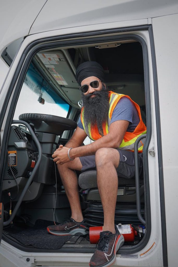 A cheerful truck driver wearing a safety vest and turban, sitting inside a vehicle and smiling.